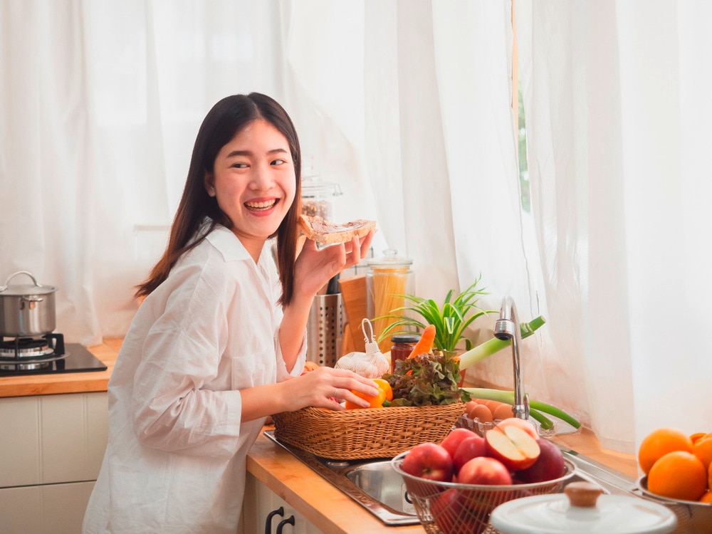 Woman eating bread while preparing vegetables and fruits in the kitchen.