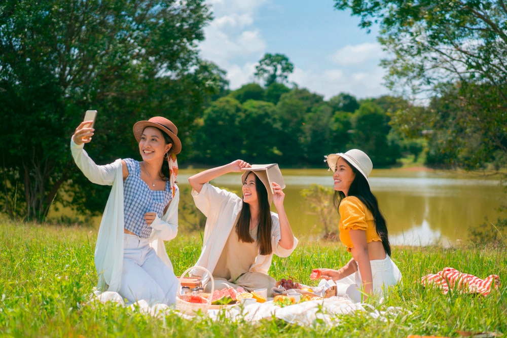 Three women taking selfies while enjoying a picnic by the lake.