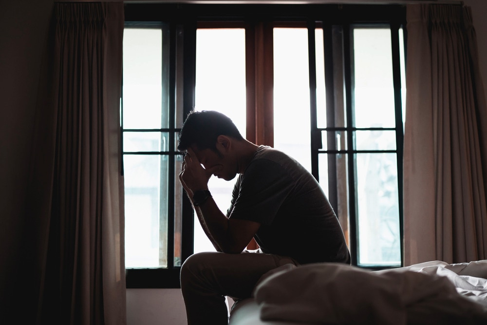 A silhouette of a man sitting on a bed holding his head in pain.