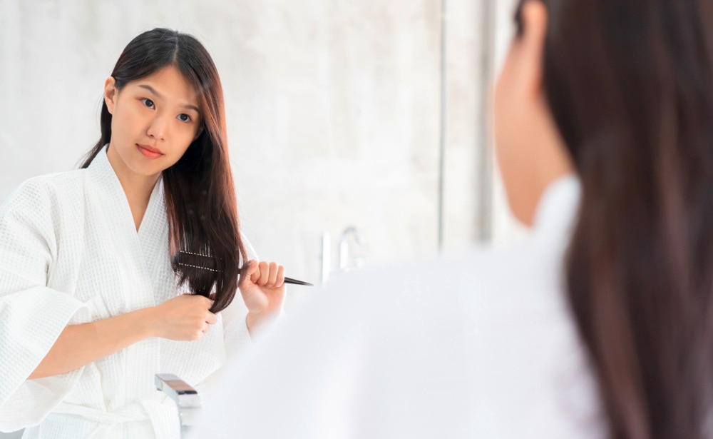 An Asian woman wearing a robe and brushing her hair in front of a mirror