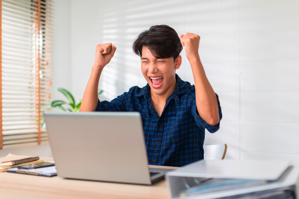 Asian man with one-length hair, rejoicing in front of a laptop.