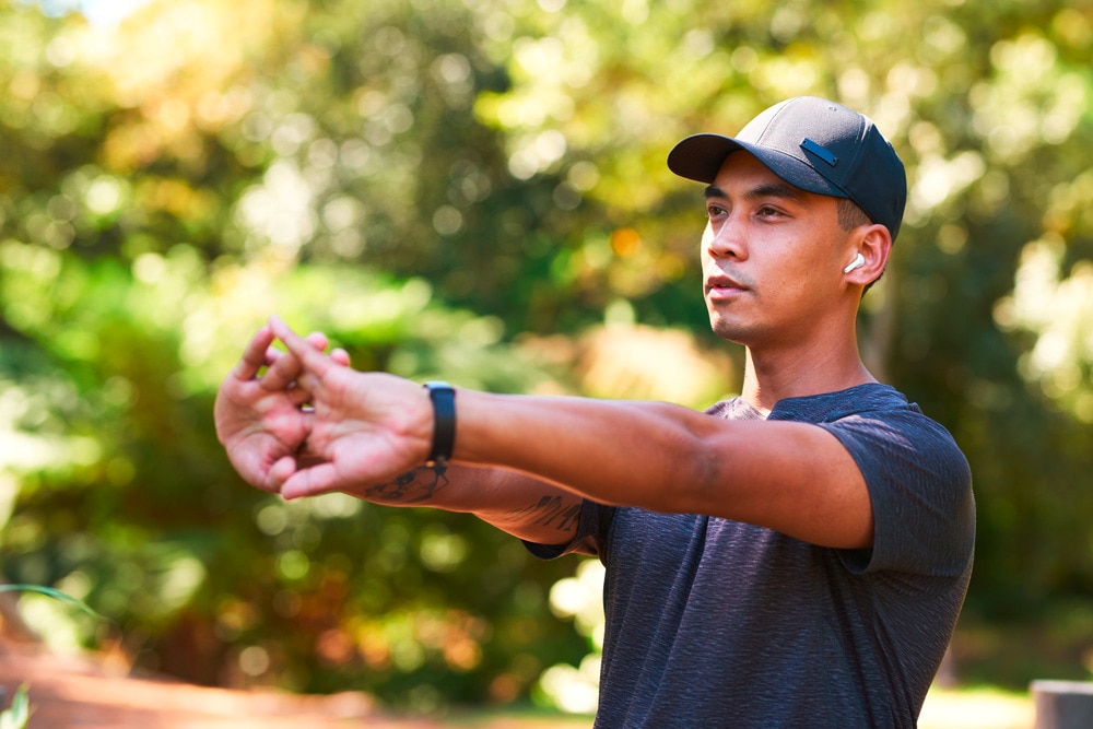 Asian man wearing a cap stretching his arms outdoors.