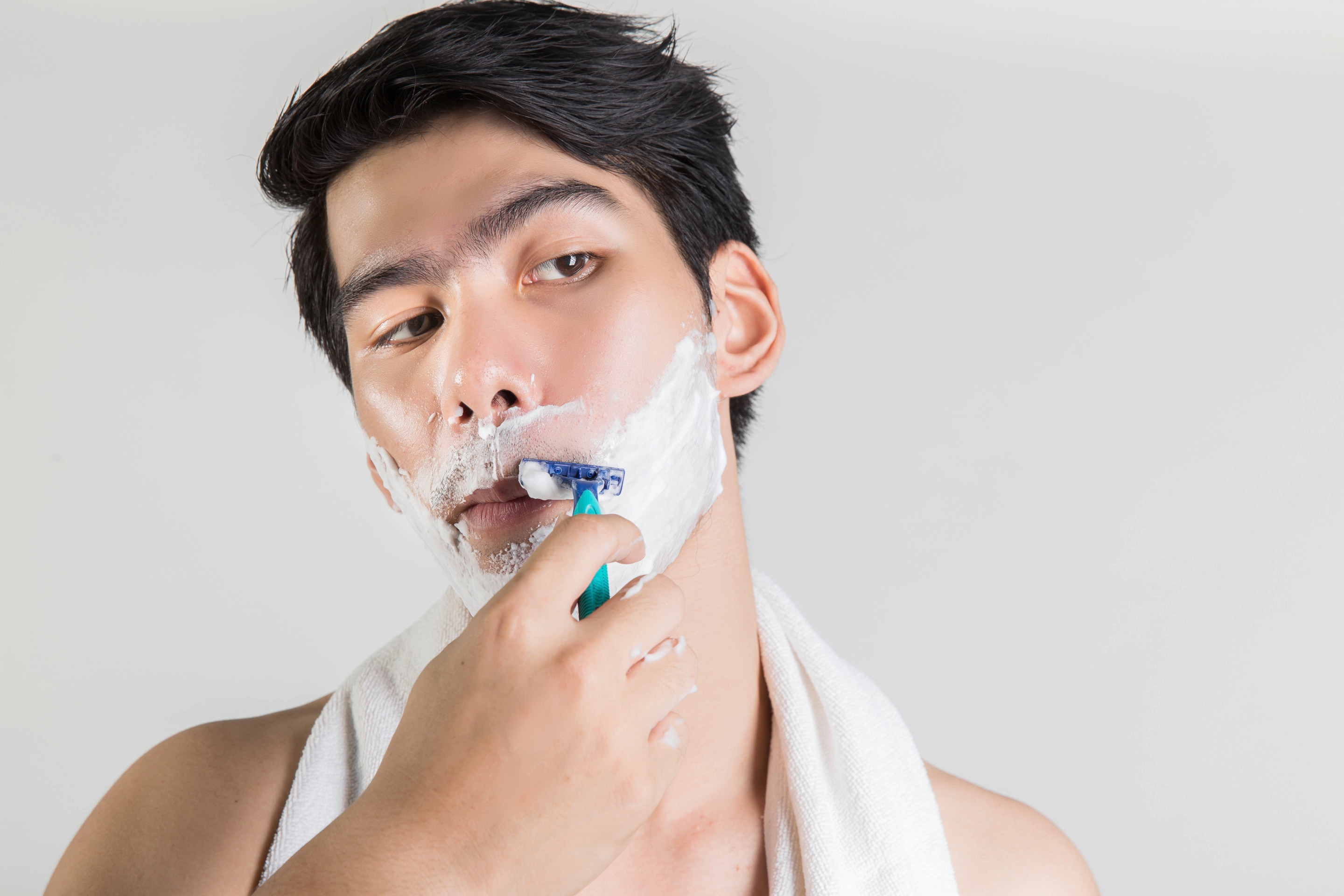 Asian man shaving with cream and a towel around his shoulders
