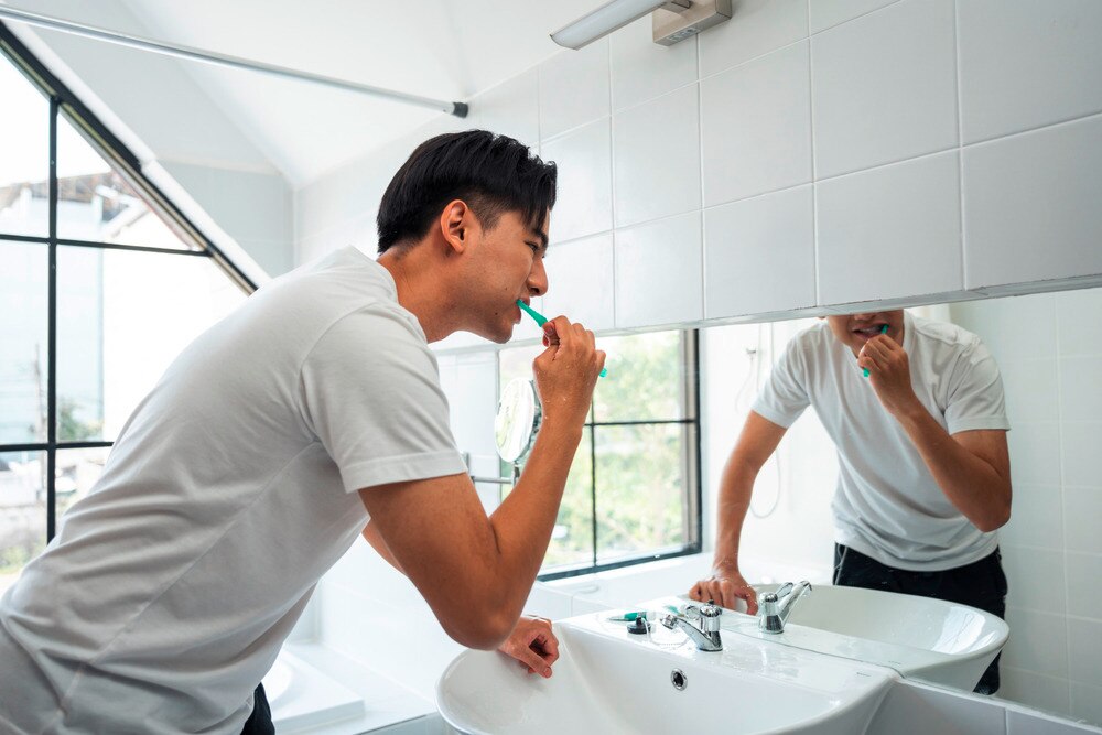 Asian man bending over sink and brushing teeth in front of mirror.