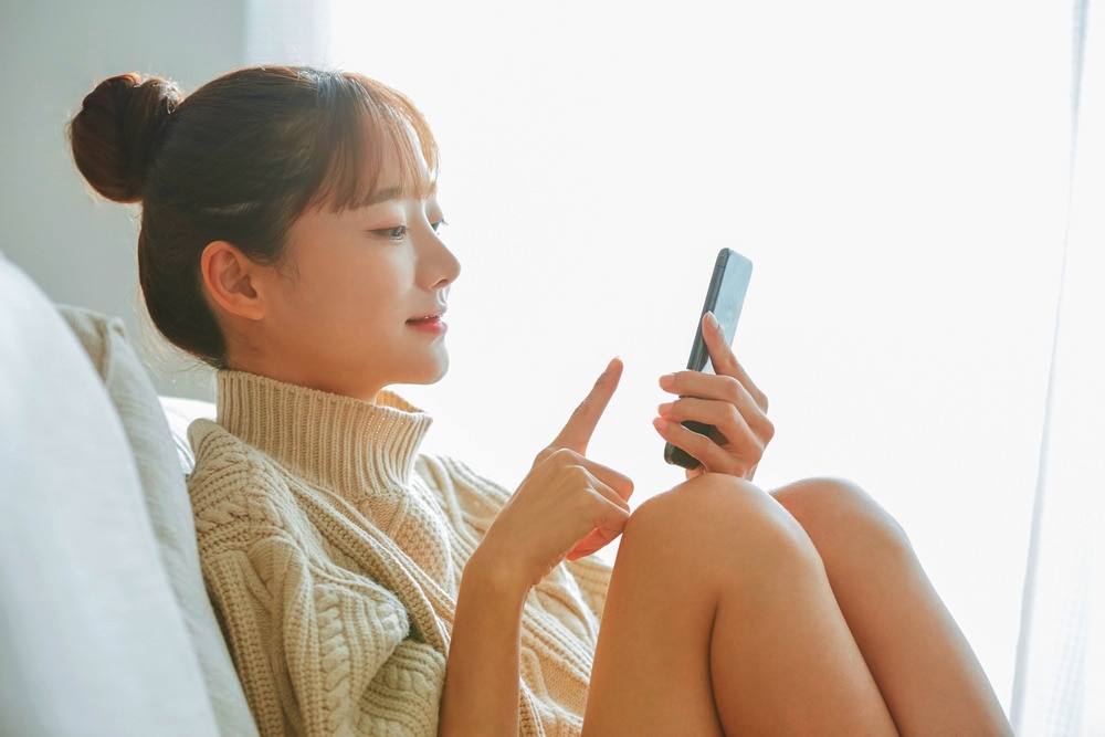 Asian woman with a top knot sitting on a sofa while browsing on her phone.