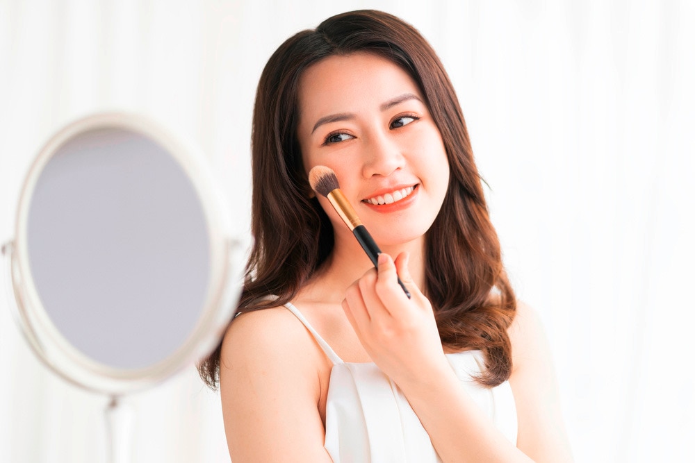 A portrait of woman applying highlighter on her cheek with a fluffy brush.