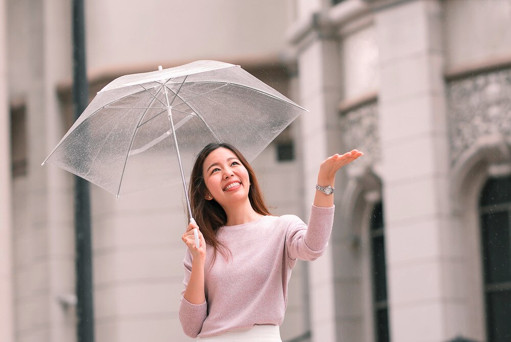 Smiling woman holding a clear umbrella and holding out her hand to catch raindrops.