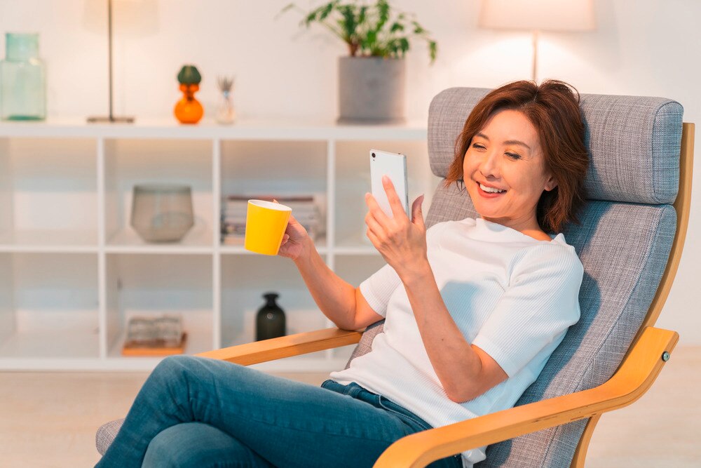 Happy Asian woman holding a yellow mug in one hand and a phone in the other.