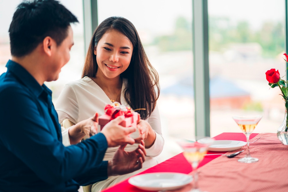 Asian man giving girlfriend a pink gift at dinner table.