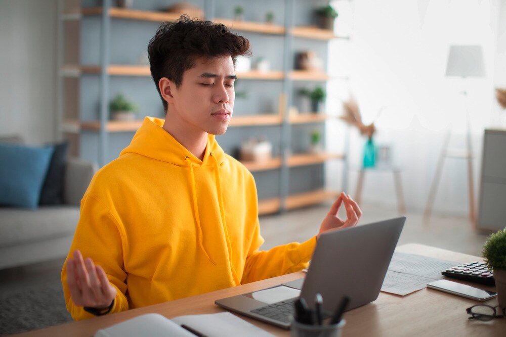 Asian man in yellow hoodie meditating in front of laptop.