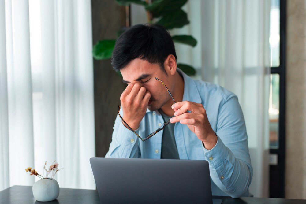 Asian man rubbing his eyes and holding glasses in front of laptop.