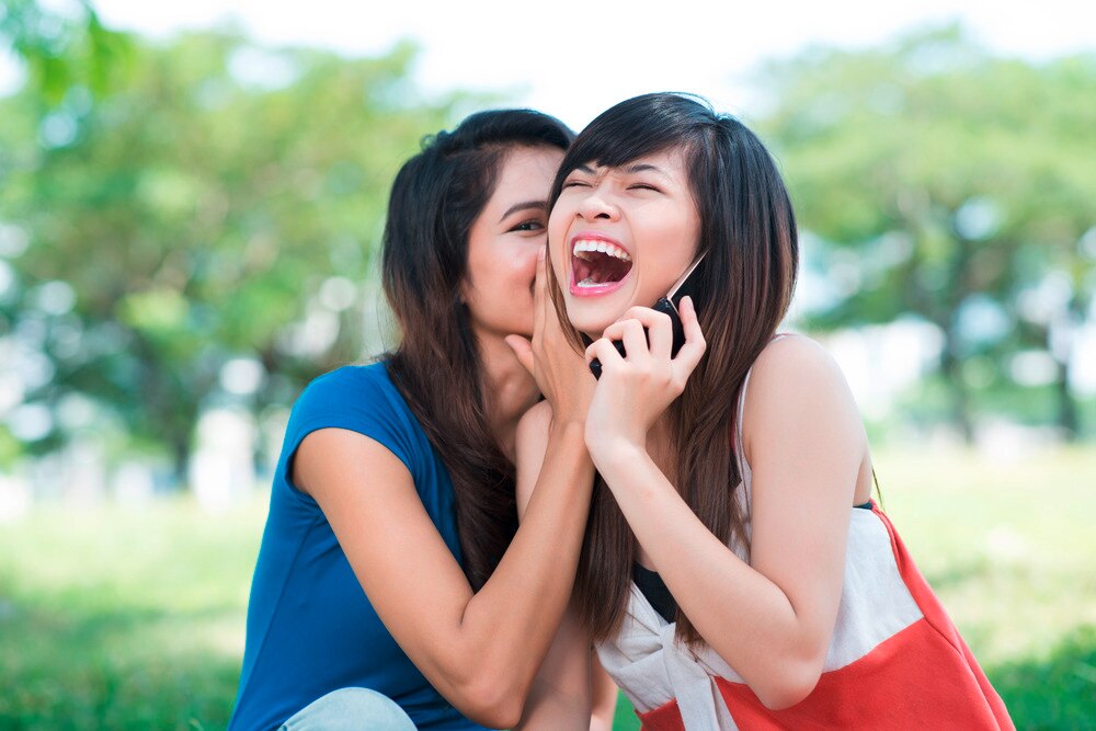 Asian friends whispering to another friend holding a cellphone while laughing.