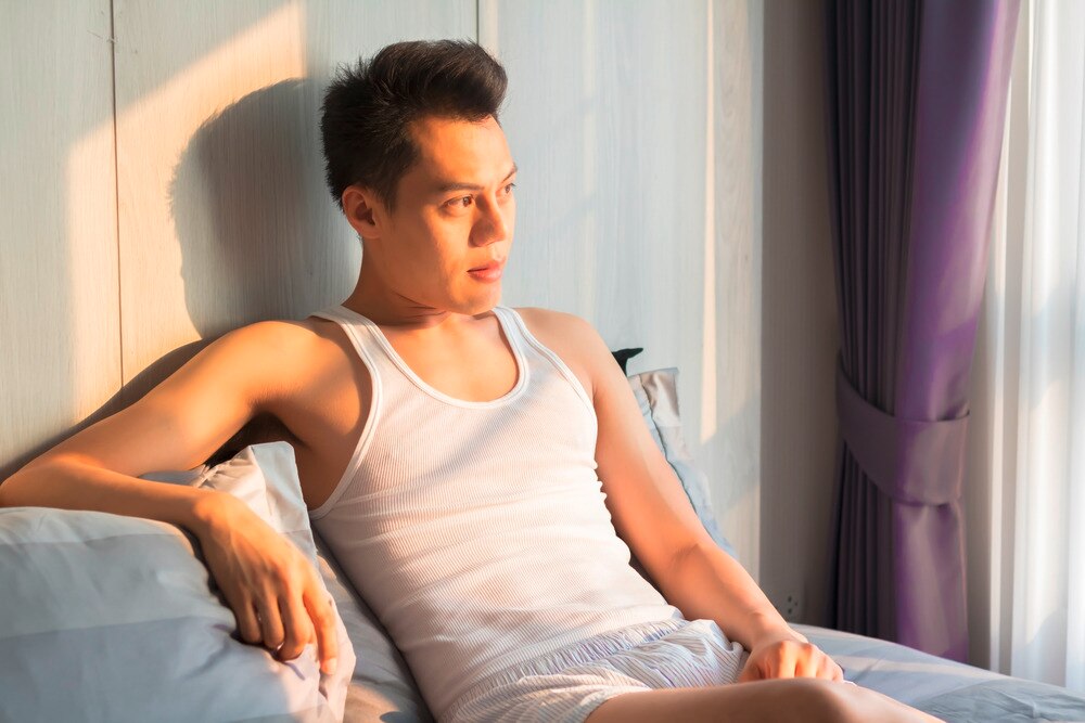 A portrait of man looking at the window while sitting on a bed.