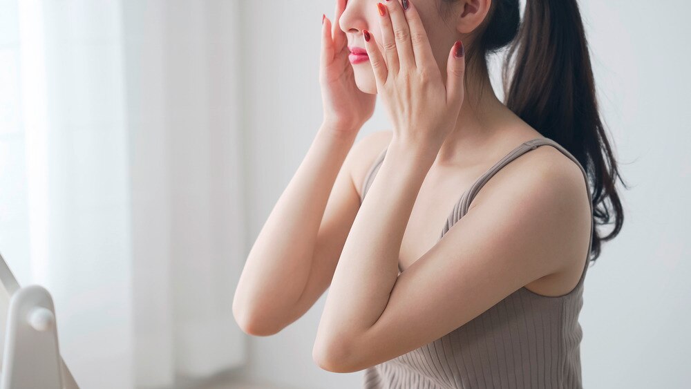 Woman with red nails applying skincare product onto her face.