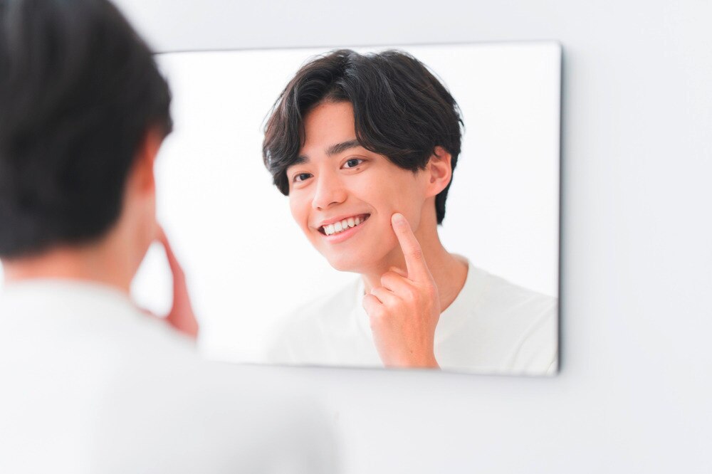 Asian man with longish hair touching face while looking in mirror.