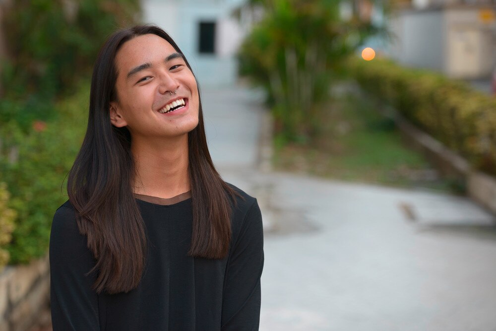 Asian man with long straight hair laughing outside.