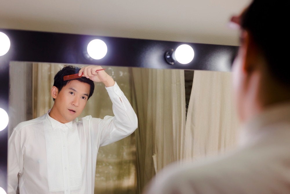 Asian man combing hair in front of mirror.