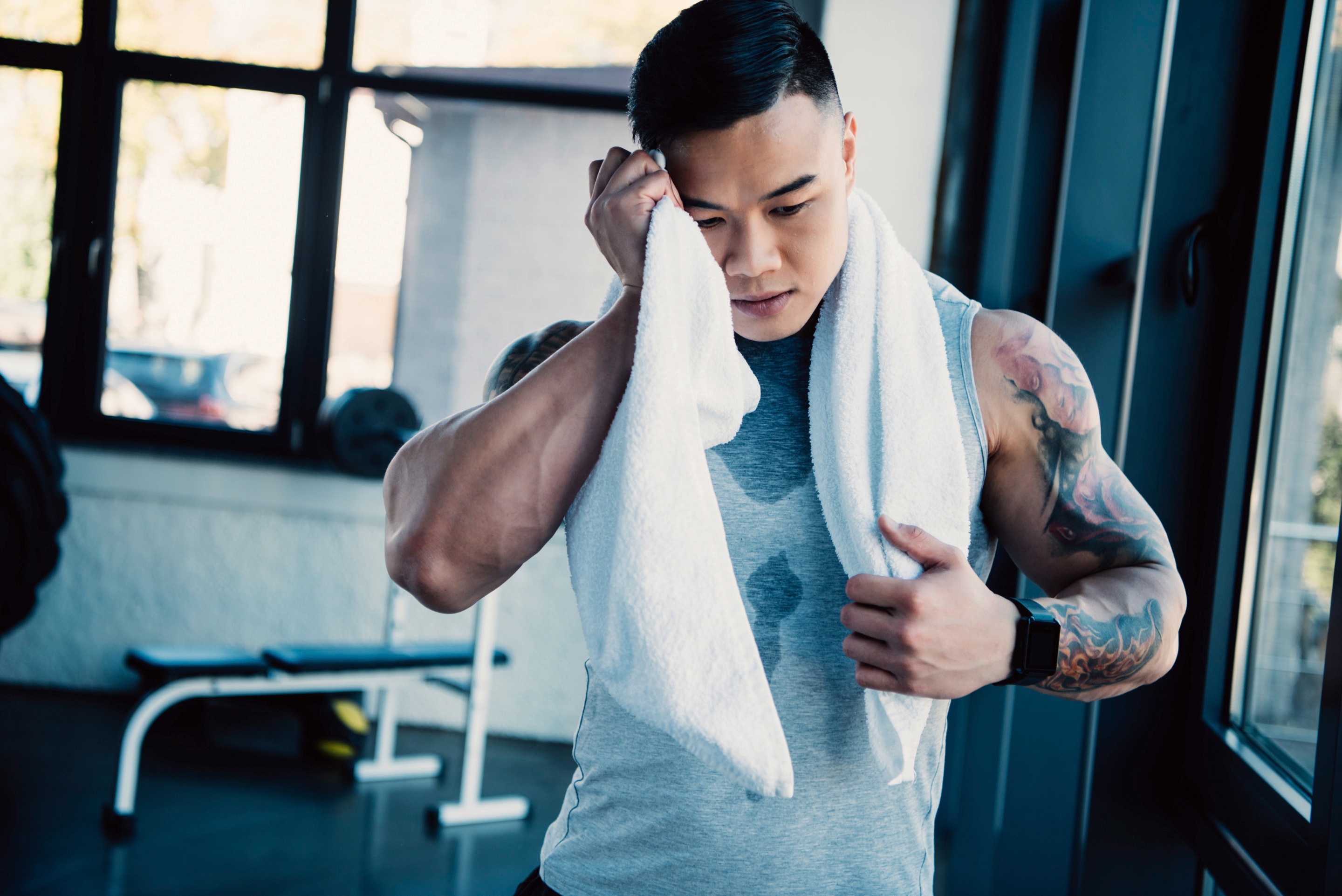 Asian man with tattoos and biceps wiping face with towel at the gym