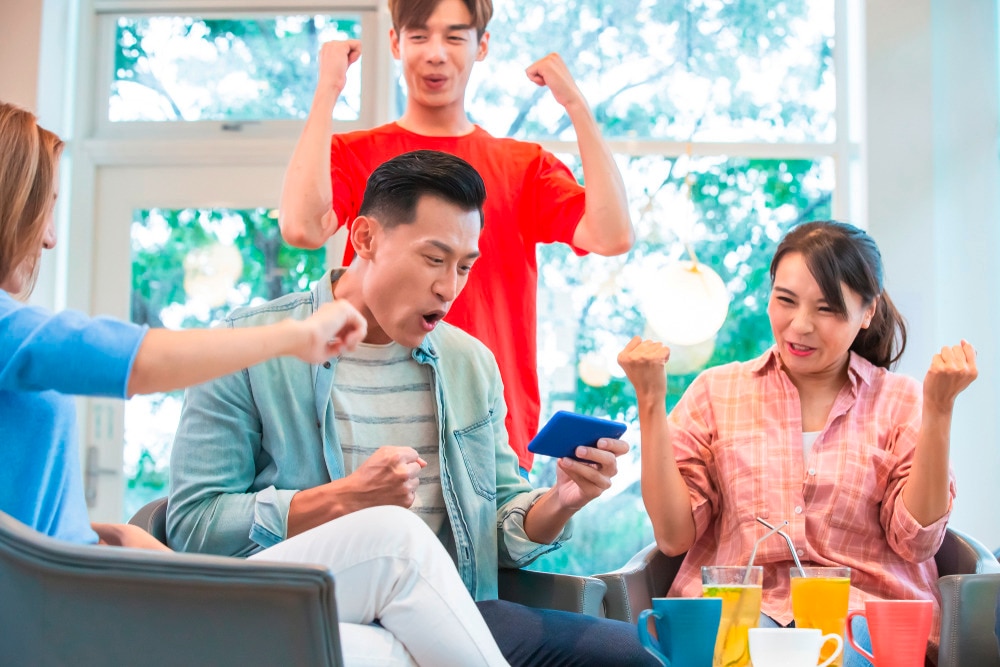 A group of friends cheering on a man who is playing a mobile game.