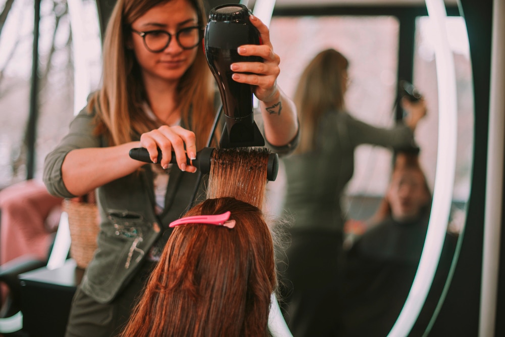 A hairdresser blow drying her client’s hair with a round brush.