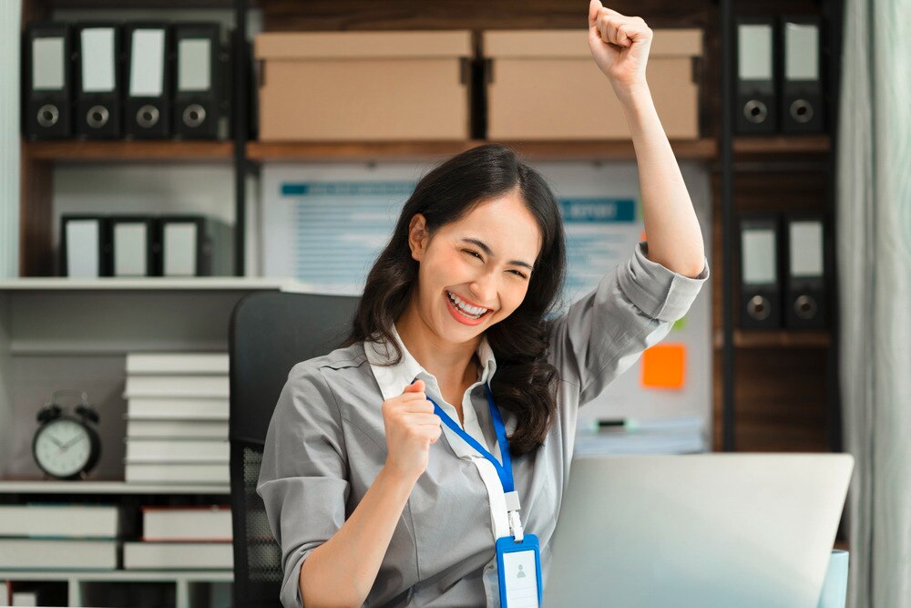 Office worker sitting in front of laptop raises her hands in joy.