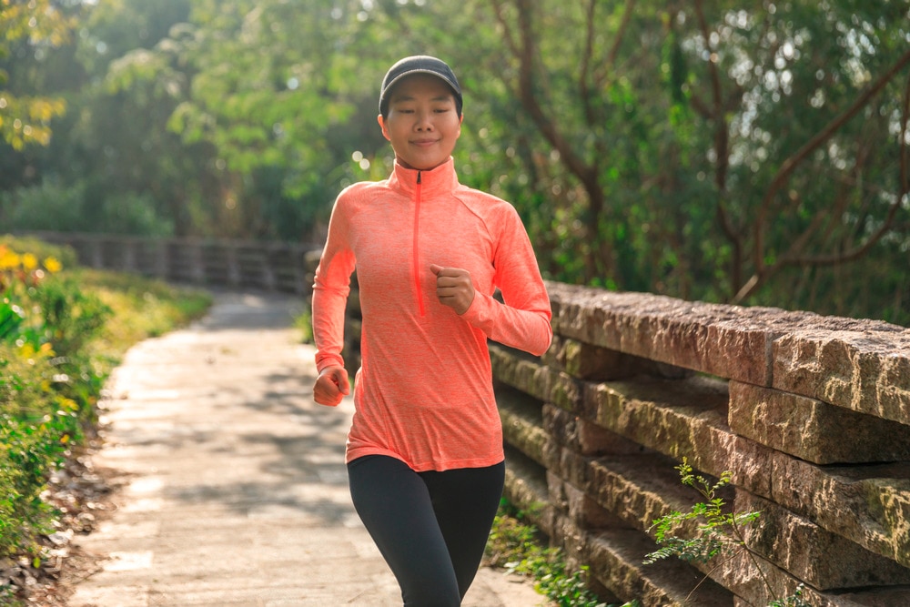 Woman in lightweight long-sleeve top, leggings, and hat jogging at the park.
