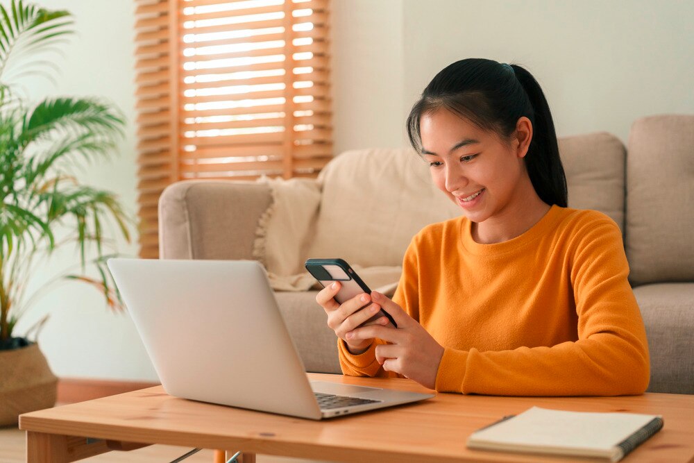 Asian girl working on laptop and holding phone at home.