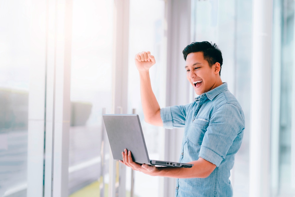 Asian male professional raises one arm in triumph as he holds a laptop in his other arm.