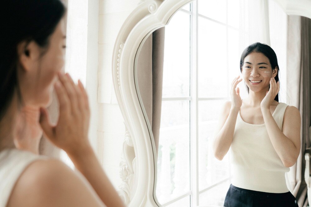 Woman applying cream onto her face while smiling in front of a mirror.