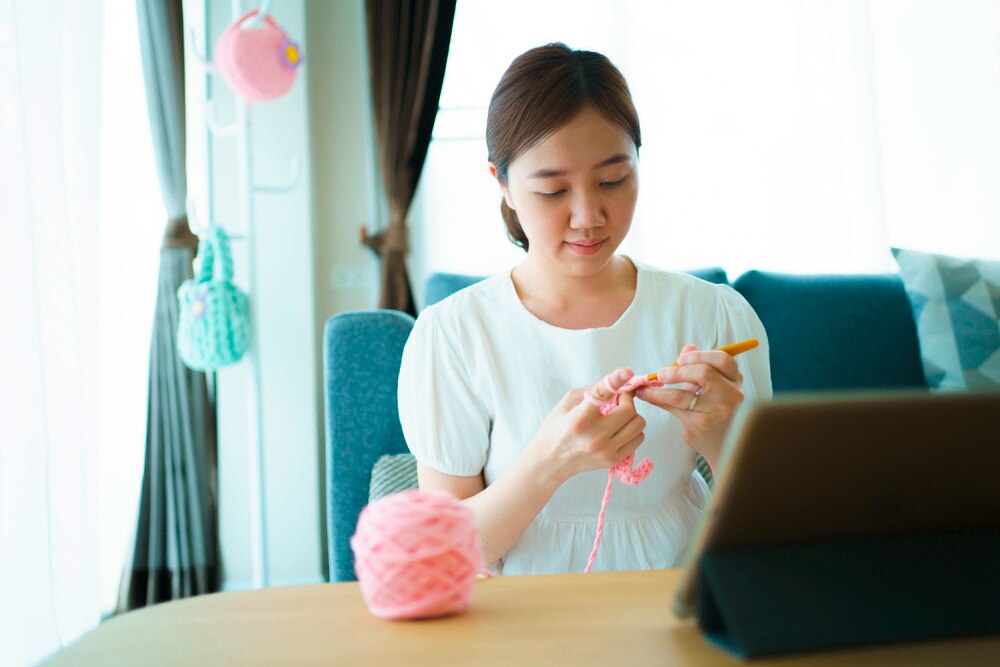 Woman crocheting a pink yarn in the living room.
