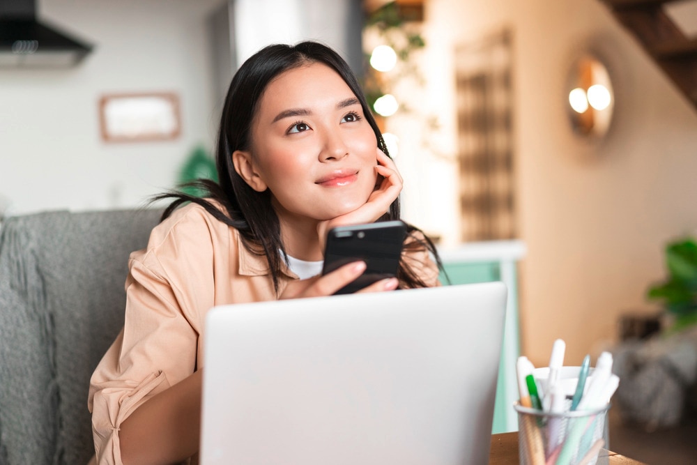 Daydreaming Asian woman in front of laptop, holding phone.