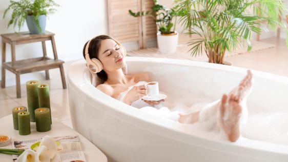 Young woman relaxing in a bathscaped bathtub with headphones on drinking a cup of tea