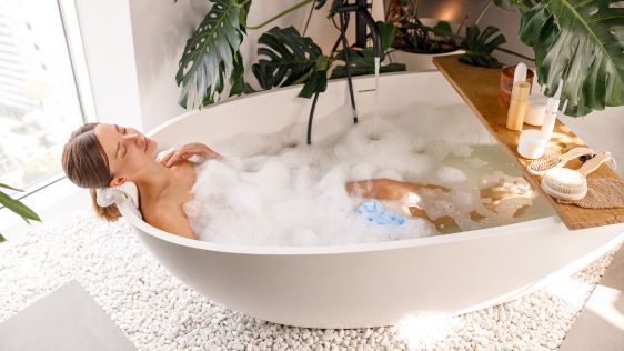 Woman relaxing in a bathscaped tub surrounded by candles and a large monstera plant 