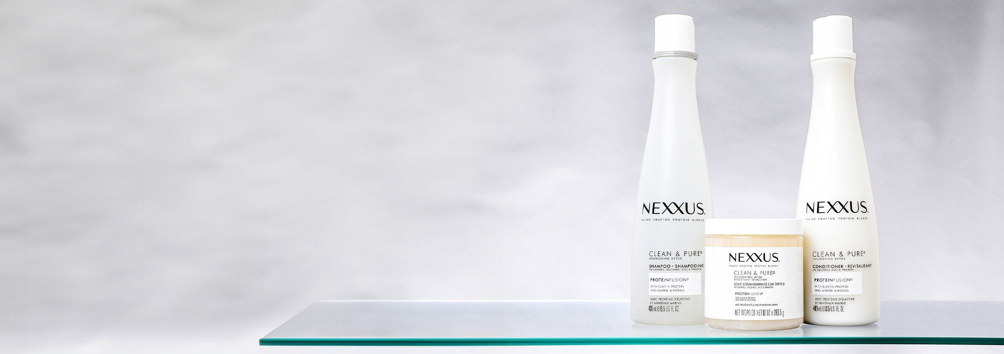 The Nexxus Clean & Pure collection, including Shampoo, Conditioner, and Scalp Scrub, is shown standing on a glass shelf against a white marble background.