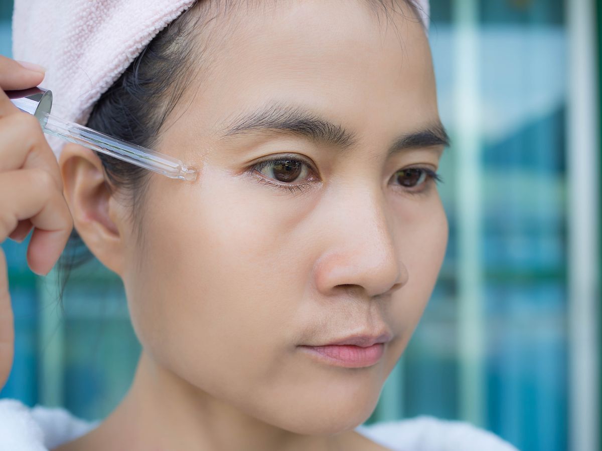 Closeup of an Asian woman applying serum on her skin using a dropper.