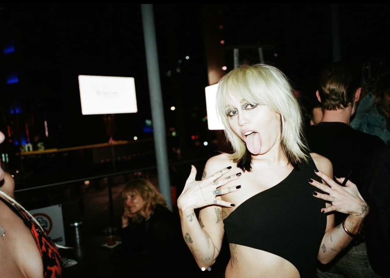 Miley Cyrus in a black crop top posing with her tongue out.