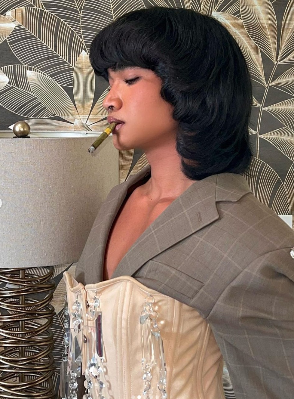 Asian with '70s style hair wearing a blazer and corset, with a cigar in their mouth.