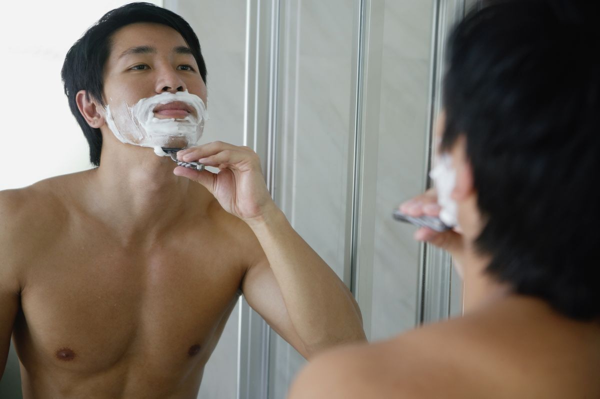 Asian man shaving in front of a mirror.