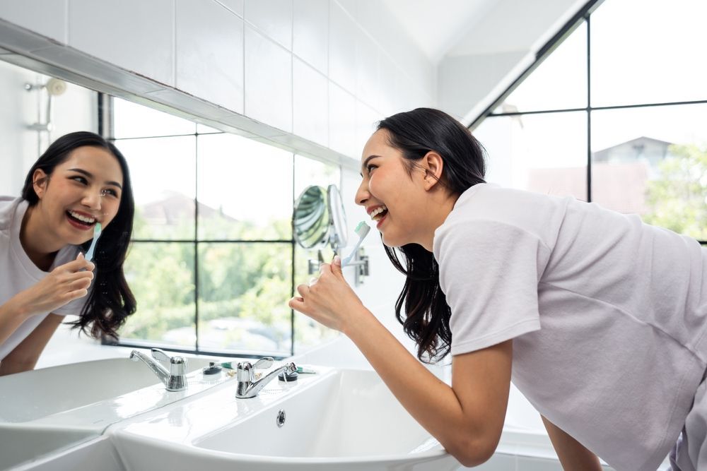Woman in white T-shirt smiling while brushing her teeth in front on the sink.