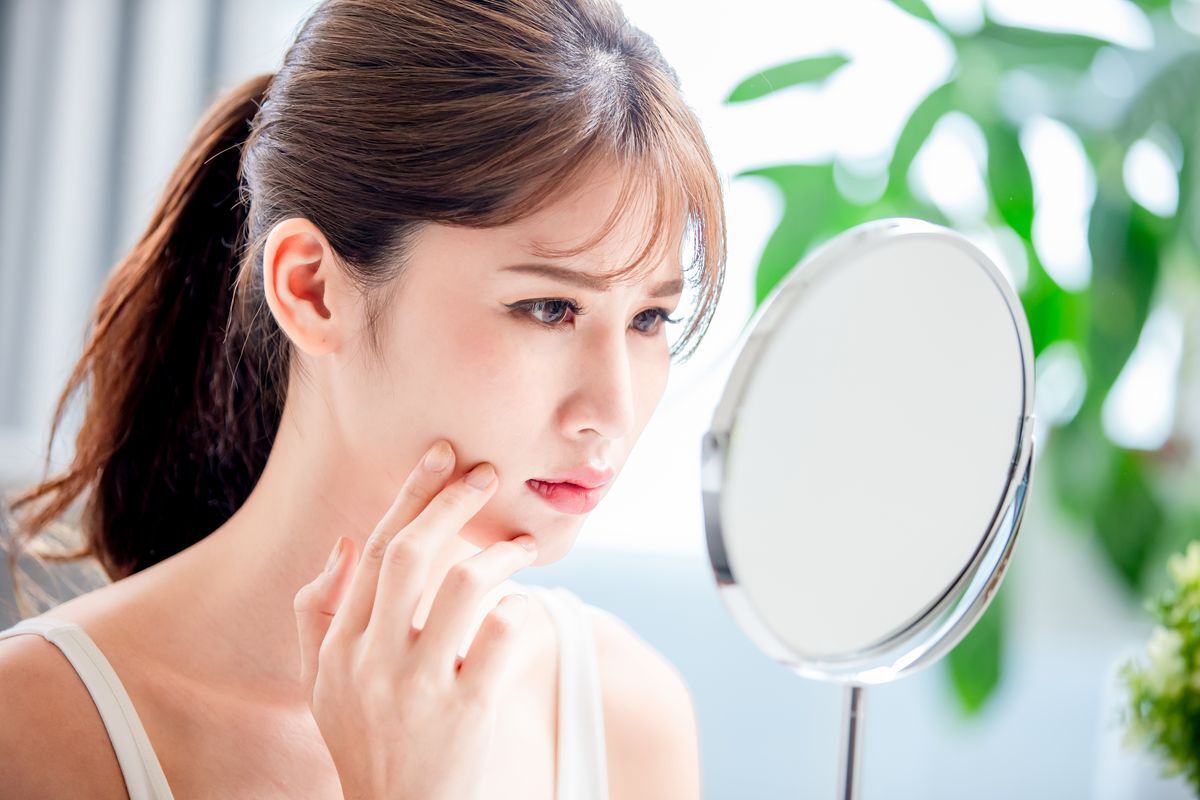 Woman in ponytail looking at her face in the mirror with a sad expression.