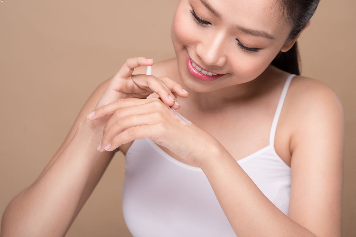 Closeup of young Asian female happily applying hand cream against a beige background.