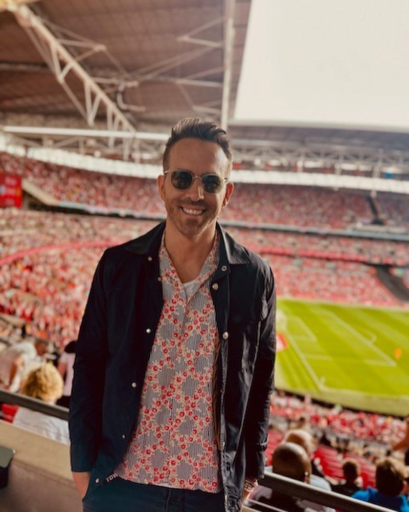 A portrait of Ryan Reynolds wearing patterned shirt and chore jacket at a sports game.