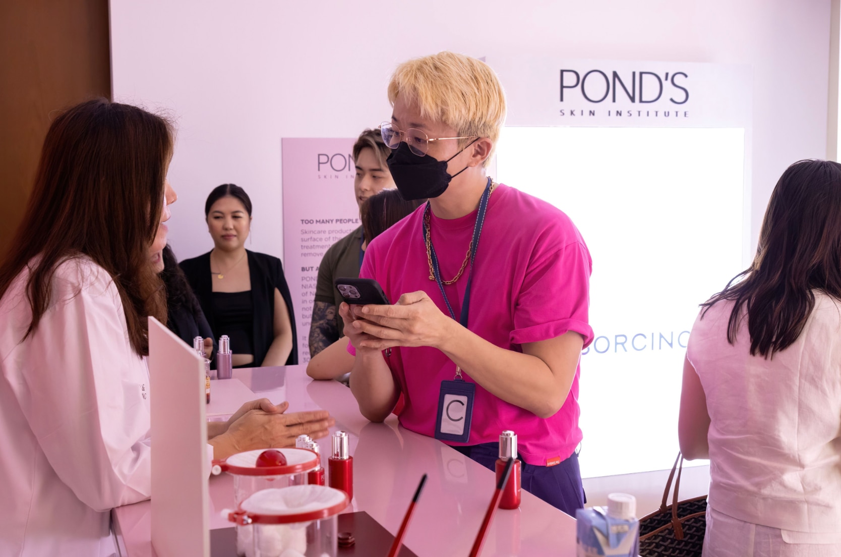 Asian man with blonde hair and pink shirt talking to a dermatologist.