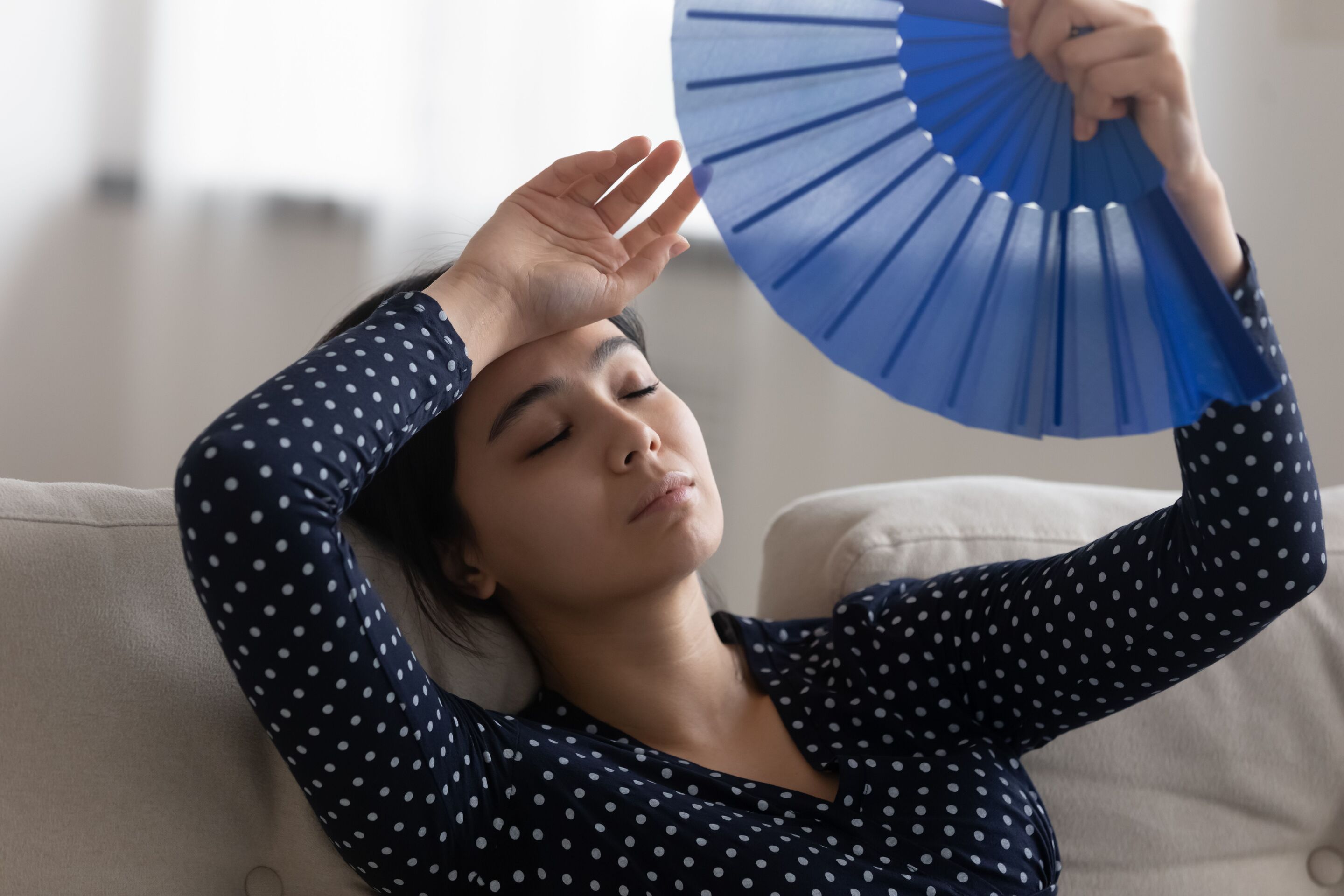 Asian woman in a polka dotted long sleeved shirt waving a fan on her face