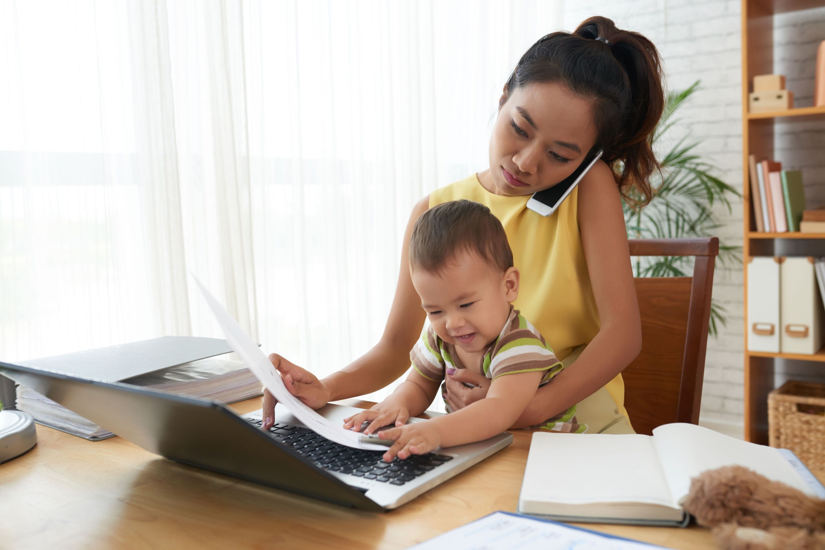 A young Asian mother working on her laptop and talking on a mobile phone while carrying a toddler