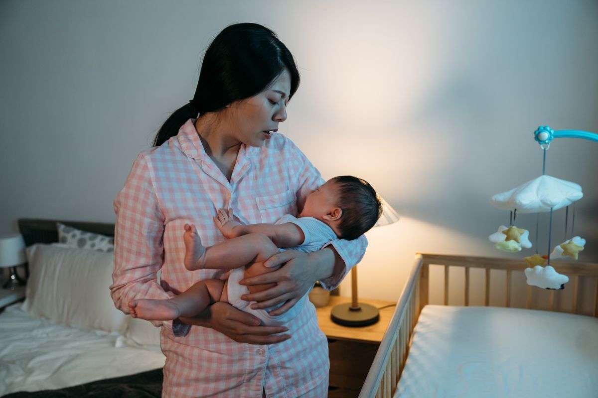 Asian mom in pink checkered pajamas carrying baby at night. 