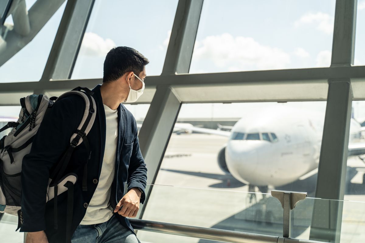 Man wearing face mask carrying backpack while looking at an airplane from the inside of an airport.