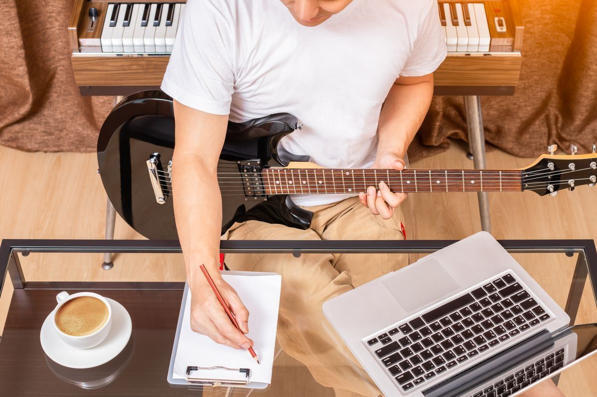 Man writing on a notepad while holding a black electric guitar.