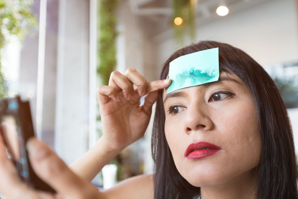 Woman using blotting paper on her forehead while looking at her reflection in a compact mirror. 