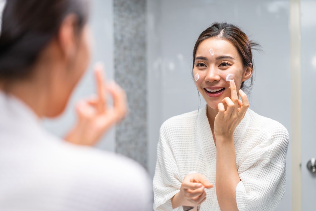 Woman in white bathrobe applying cleansing balm on her face. 